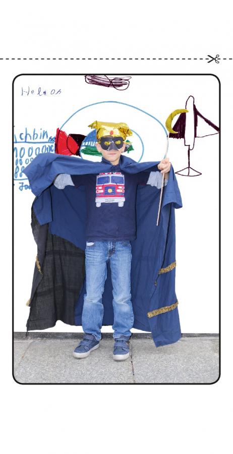 Ein Kind trägt ein Superheldenkostüm wie Superman, hinter ihm eine Wand mit einigen Zeichnungen.