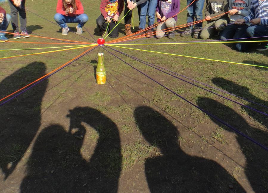 Eine Gruppe von Kindern steht im Kreis, jeder hält ein Seil, die Seile treffen sich in der Mitte, darauf liegt ein Apfel und darunter eine Saftflasche.