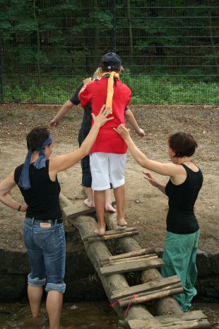 Ein kleiner Wassergraben mit einer kleinen Holzbrücke, über die zwei Kinder gehen, während zwei Frauen am Wasser stehen und den Kindern beim Überqueren helfen.